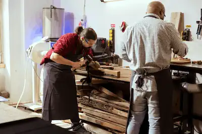 Twee personen werken in een timmerwerkplaats. De ene is hout aan het bewerken op een werktafel, terwijl de andere, met de rug naar de camera, lijkt een gereedschap aan te passen.