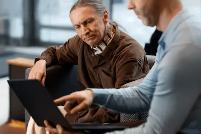 Een senior ontvangt gepersonaliseerde IT-hulp thuis, een expert helpt hem navigeren op een laptop.